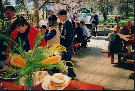 A group of people are scattered across two wooden tables, with more standing at a table in the front of the photograph where someone is serving themselves food, and others are talking. There are two men in the background playing what appears to be table soccer, with a row of cars parked behind them. The area looks to be an outdoor undercover dining area. 