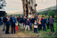 A group of over twenty people are standing outside next to a field. There is a bus to the left of the group, a massive tree behind the group, and rolling hills in the background. Most of the people pictured are facing the camera, but some are facing the back. 