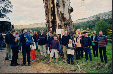 A group of over twenty people are standing outside next to a field. There is a bus to the left of the group, a massive tree behind the group, and rolling hills in the background. Most of the people pictured are facing the camera, but some are facing the back. 