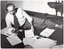 B&W photo of a man, wearing a shirt, tie, and trousers, is lying on his side across a carpeted floor. He is propped up on one elbow, and is holding a small recording device or dictaphone in one hand. In his other hand, he is holding some papers. On the floor in front of him are several papers, an envelope, folders, and other printed material. There is a bunch of keys on one of the papers, with a box of Redheads matches and a pen beside them. Behind him is an office/desk chair on castors and a mug on the floor.