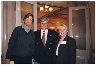 COL photo of three people standing close together, in a row. They are in front of an arched doorway with double doors, which are open. Each door has a wooden panel and etched glass.