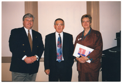 COL photo of three people standing close together in a large hall. One person is holding an information folder.