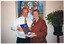 COL photo of two people standing close to one another. They are each holding some publications, one of which is titled 'Diversity Action Plan'. Behind them is a large painting on the wall, and beside them is a large potted indoor plant on a wooden stand.