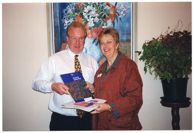 COL photo of two people standing close to one another. They are each holding some publications, one of which is titled 'Diversity Action Plan'. Behind them is a large painting on the wall, and beside them is a large potted indoor plant on a wooden stand.