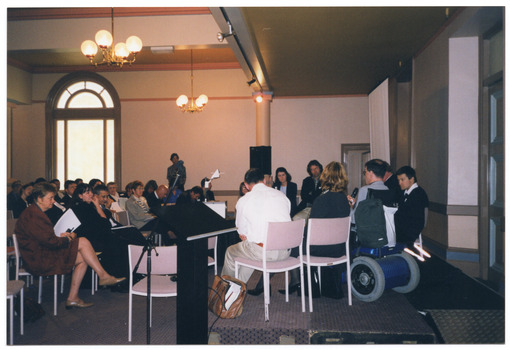 COL photo of several people seated on a small stage (their backs to the camera), facing an audience, who are sitting on rows of chairs. They are in a large hall. One of the presenters is a wheelchair user. 