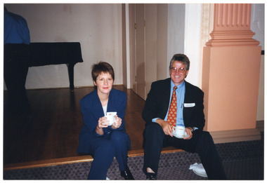 COL photo of two people sitting on the edge of a small stage, holding cups of tea.