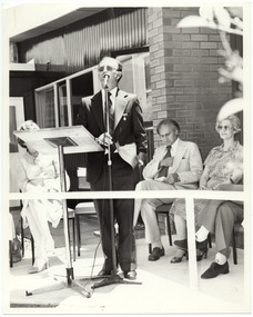 B&W photo of a man standing at a podium on the veranda of a building. He is speaking into a microphone on a stand.  There are several people seated behind him, three of whom are visible within the frame, while two others are partially obscured.