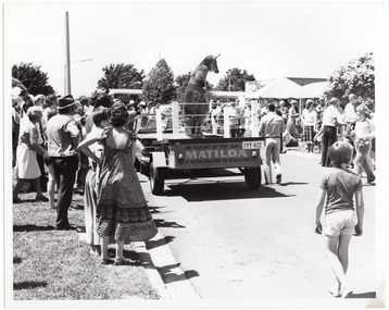 B&W photo of a crowd gathered around a car and trailer (with the registration number IYY-422). A person dressed in a kangaroo costume and boxing gloves is standing on the trailor. On the rear panel of the trailor, there is a wooden board with the text "The film hit of the year… MATILDA See Matilda challenge the world champ". In the background, there are some market stalls.