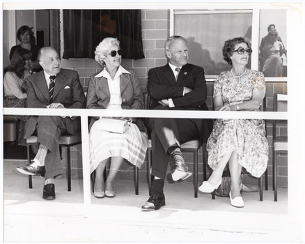 B&W photo of four people sitting on chairs, on the veranda of a small building. Behind them and to their right, two other people can be seen near a doorway (one is standing, while the other is seated). On the left, some people's reflections are visible in the window.