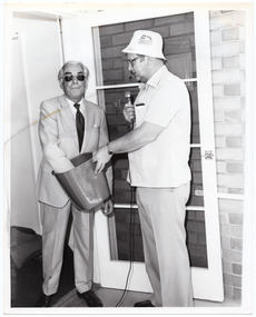 B&W photo of two people standing in an open doorway. One of them is wearing a suit and tie, and sunglasses. The other is wearing a fabric hat with "3MP Radio" branding and eye glasses. He is holding a microphone and a plastic waste paper basket while the man in the suit has one arm inside the basket. 