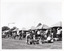 B&W photo of various market stalls set up under a row of gazebos, with vendors and people browsing. In the foreground, a man is looking down at a group of boxes.