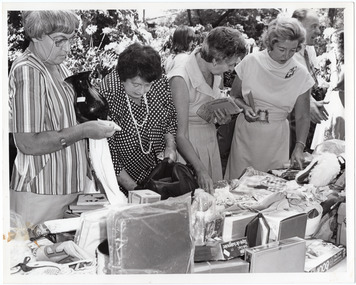 B&W photo of four women standing in front of a trade stall table, perusing the goods for sale. There are several other people in the background.