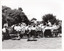 B&W photo of a brass band playing on a grassy area outdoors. A conductor is leading the band. On the drummer's drum, the text "Kew Citize" (partially obscured) is visible. In the background, there are people browsing amongst market stall tents.