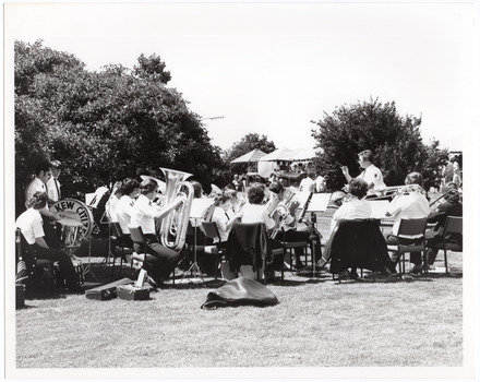 B&W photo of a brass band playing on a grassy area outdoors. A conductor is leading the band. On the drummer's drum, the text "Kew Citize" (partially obscured) is visible. In the background, there are people browsing amongst market stall tents.