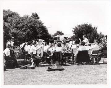 B&W photo of a brass band playing on a grassy area outdoors. A conductor is leading the band. On the drummer's drum, the text "Kew Citize" (partially obscured) is visible. In the background, there are people browsing amongst market stall tents.