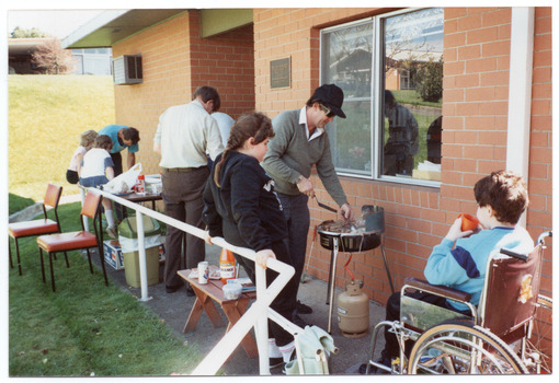 COL photo of a few adults and children gathered outside a brick building. One man is cooking steaks on a portable barbeque, while another is setting up a table with various food items. A few children are watching the adults setting up. One child is a wheelchair user. He has his left side and back to the camera, in three-quarter profile.