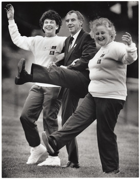 B&W photo of three people taking part in an exercise demonstration outdoors. A man, standing between two women, kicks one leg high in the air. The women, standing on either side of him, link their arms into his as each one holds her other arm out to the side. One of the women is also kicking her leg forward.
