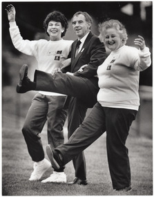 B&W photo of three people taking part in an exercise demonstration outdoors. A man, standing between two women, kicks one leg high in the air. The women, standing on either side of him, link their arms into his as each one holds her other arm out to the side. One of the women is also kicking her leg forward.