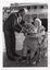 B&W photo of a man presenting a woman with a large bouquet of flowers. The man, who is standing, leans forward towards the woman, who is a wheelchair user. They are located on a paved outdoor area. In the background, there is a large two-storey house which is located on slightly higher ground.
