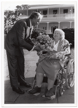 B&W photo of a man presenting a woman with a large bouquet of flowers. The man, who is standing, leans forward towards the woman, who is a wheelchair user. They are located on a paved outdoor area. In the background, there is a large two-storey house which is located on slightly higher ground.