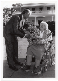 B&W photo of a man presenting a woman with a large bouquet of flowers. The man, who is standing, leans forward towards the woman, who is a wheelchair user. They are located on a paved outdoor area. In the background, there is a large two-storey house which is located on slightly higher ground.