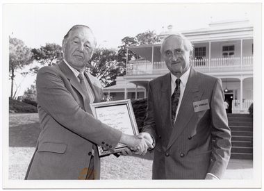 B&W photo of two men shaking hands while one of them holds a framed certificate in his other hand. They are standing in a large garden. In the background, there is a large two-storey house, which is located on slightly higher ground. A woman (partially obscured) is standing in front of the house.