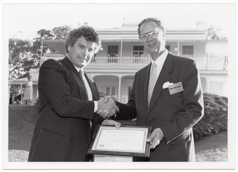 B&W photo of two men holding a framed certificate while shaking hands. They are standing in a large garden. In the background, there is a large two-storey homestead, which is located on slightly higher ground. A woman standing in front of the house, to the left of the frame.