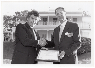 B&W photo of two men holding a framed certificate while shaking hands. They are standing in a large garden. In the background, there is a large two-storey homestead, which is located on slightly higher ground. A woman standing in front of the house, to the left of the frame.