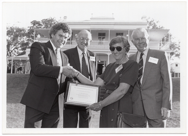 B&W photo of four people posing for a photo on the lawns of an historic home. A man is shaking the hand of a woman, who is holding a framed certificate, while two other men look on. They are standing in a large garden. In the background, there is a large two-storey house, which is located on slightly higher ground. There is a woman (partially obscured) standing in front of the house, to the left of the frame.
