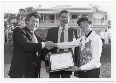 B&W photo of a man shaking hands with a woman while another man, standing between them, looks on. They are standing in a large garden. In the background, there is a large two-storey house, which is located on slightly higher ground.