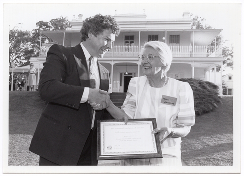 B&W photo of a man shaking hands with a woman while they both hold a framed certificate. They are standing in a large garden. In the background, there is a large two-storey house, which is located on slightly higher ground. A woman is standing in front of the house, to the left of the frame.