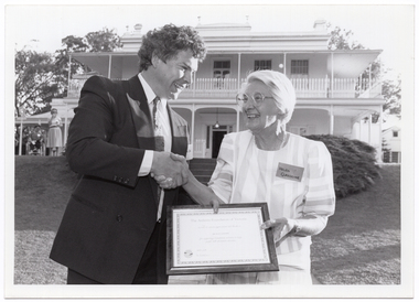 B&W photo of a man shaking hands with a woman while they both hold a framed certificate. They are standing in a large garden. In the background, there is a large two-storey house, which is located on slightly higher ground. A woman is standing in front of the house, to the left of the frame.