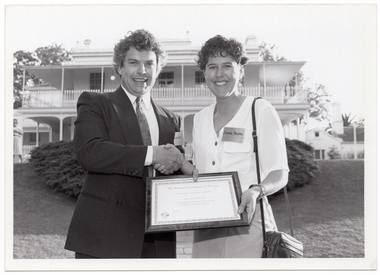 B&W photo of a man shaking hands with a woman while they both hold a framed certificate. They are standing in a large garden. In the background, there is a large two-storey house, which is located on slightly higher ground. There is a woman standing in front of the house, to the left of the frame.