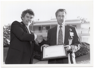 B&W photo of two men shaking hands while holding a framed certificate. They are standing in a large garden. In the background, there is a large two-storey house, which is located on slightly higher ground.