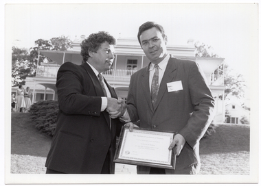 B&W photo of two men shaking hands while holding a framed certificate. They are standing in a large garden. In the background, there is a large two-storey house, which is located on slightly higher ground. Two women are standing in front of the house, to the left of the frame.