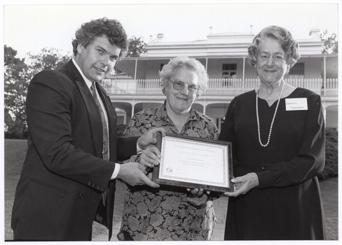 B&W photo of a man and two women holding a framed certificate. They are standing in a large garden. In the background, there is a large two-storey house, which is located on slightly higher ground.
