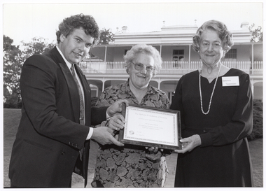 B&W photo of a man and two women holding a framed certificate. They are standing in a large garden. In the background, there is a large two-storey house, which is located on slightly higher ground.
