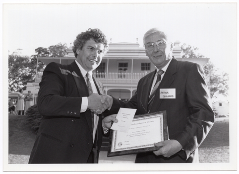 B&W photo of two men shaking hands while holding a framed certificate. One of the men also has a card and envelope in his hand. They are standing in a large garden. In the background, there is a large two-storey house, which is located on slightly higher ground. Two women are standing in front of the house, to the left of the frame.