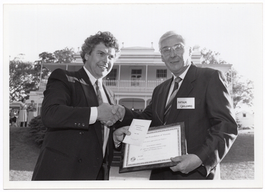 B&W photo of two men shaking hands while holding a framed certificate. One of the men also has a card and envelope in his hand. They are standing in a large garden. In the background, there is a large two-storey house, which is located on slightly higher ground. Two women are standing in front of the house, to the left of the frame.