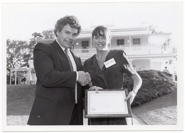 B&W photo of a man and woman shaking hands while holding a framed certificate. They are standing in a large garden. In the background, there is a large two-storey house, which is located on slightly higher ground. Two women standing in front of the house, to the left of the frame.