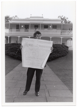 B&W photo of a man standing in an outdoor paved area in front of a large two-storey house. He is holding up a large building plan.