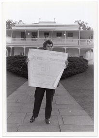 B&W photo of a man standing in an outdoor paved area in front of a large two-storey house. He is holding up a large building plan.