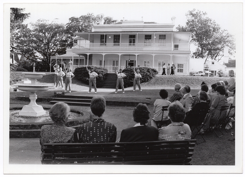 B&W photo of a group of six women conducting an exercise demonstration. They are on a paved and grassy area of a large garden, in front a large two-storey house. In the foreground, there are several people sitting on rows of chairs and a park bench, beside a large fountain. In the background, there are several people standing in front of the house - one woman to the left of the frame and four women to the right of the frame.
