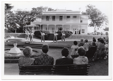 B&W photo of a group of six women conducting an exercise demonstration. They are on a paved and grassy area of a large garden, in front a large two-storey house. In the foreground, there are several people sitting on rows of chairs and a park bench, beside a large fountain. In the background, there are several people standing in front of the house - one woman to the left of the frame and four women to the right of the frame.