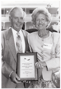 B&W photo of a man and a woman holding a framed award certificate. They are standing on the grounds of a large two-storey house, which is in the background.