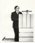B&W photo of a man standing at a wooden podium on a stage.