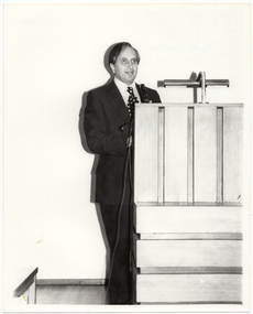 B&W photo of a man standing at a wooden podium on a stage.