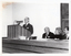 B&W photo of a man standing at a podium. The podium is built into a large wooden desk, behind which are seated three other people.