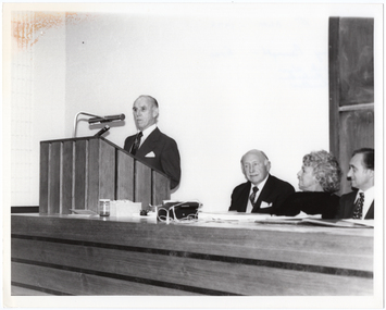 B&W photo of a man standing at a podium. The podium is built into a large wooden desk, behind which are seated three other people.