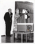 B&W photo of a woman standing at a tall wooden podium. There is a man standing behind her. Beside the podium is a large slide projector on a table.